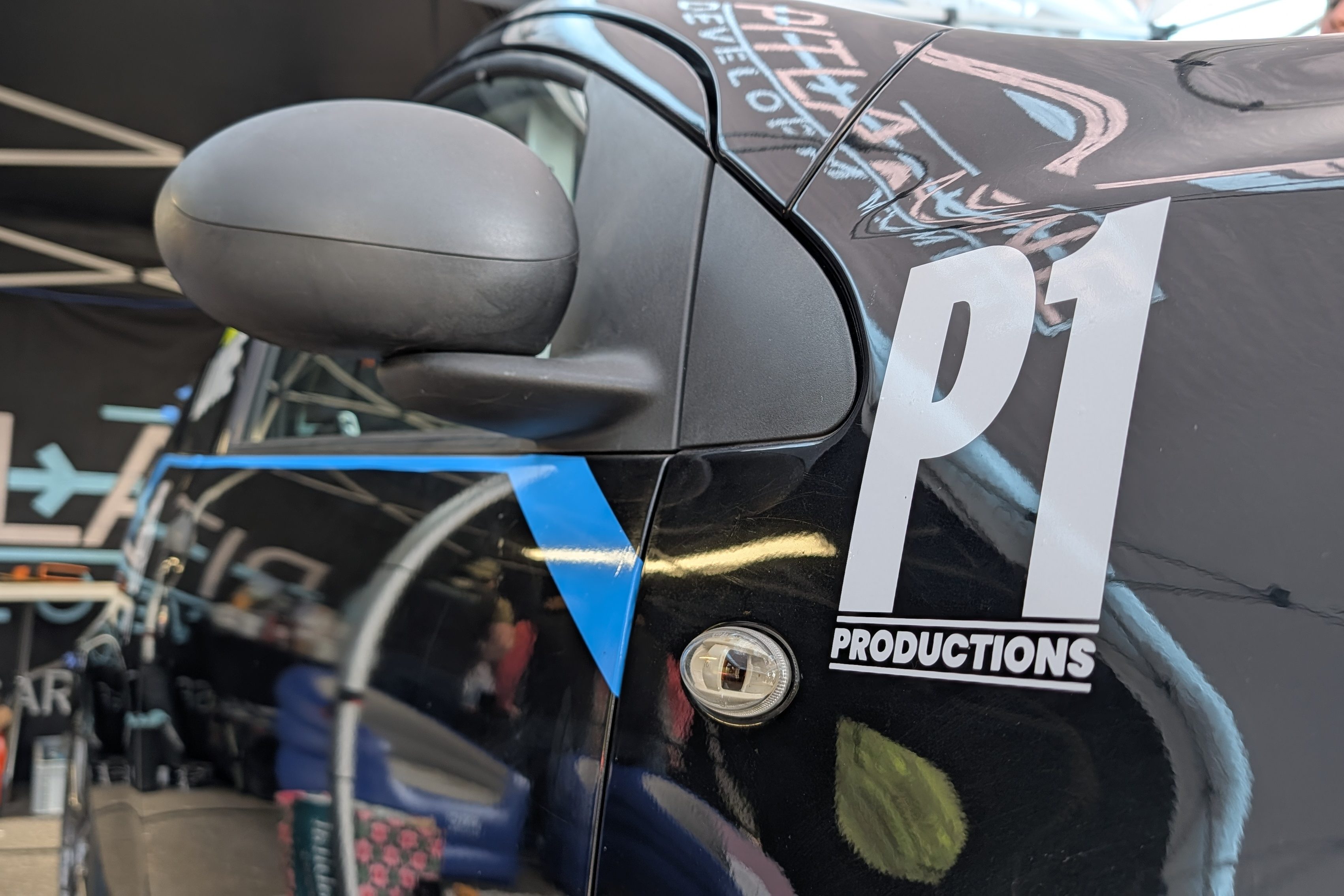 Close-up view of a black car door with a side mirror, featuring a 'P1 PRODUCTIONS' sticker and a blue design element.